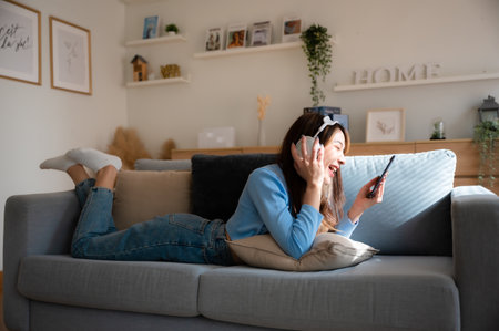 Happy young asian woman relaxing at home. Female smile sitting on sofa and holding mobile smartphone. Girl using video call to friend. Listening to musicの写真素材
