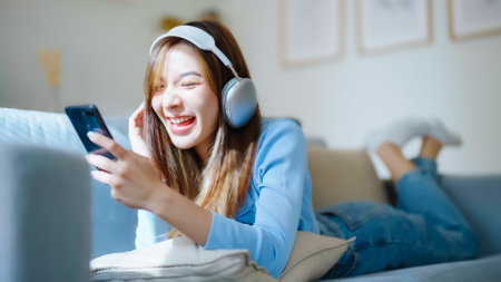 Happy young asian woman relaxing at home. Female smile sitting on sofa and holding mobile smartphone. Girl using video call to friend. Listening to musicの写真素材