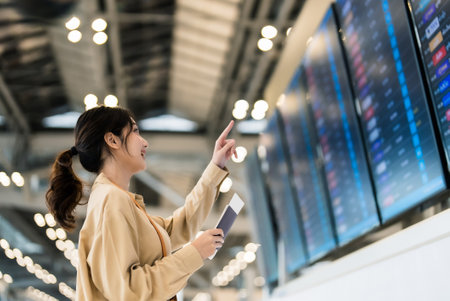 Young asian woman with passport and boarding pass as a hand in international airport looking at the flight information board, checking her flightの写真素材