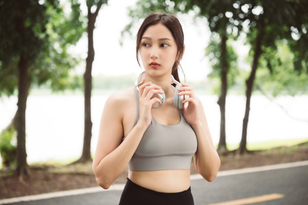 Asian woman trainer is engaged in fitness in public park. Female wearing sport bra and using headphone listening to music. Healthy lifestyle, sports outdoor activities in parkの写真素材