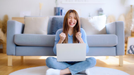 Young asian woman in good spirits working on laptop at home while sitting on the floor close to the couch. Excited female winner celebrating success while glancing at notebookの写真素材