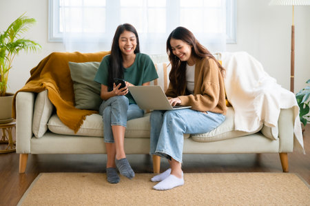 Two asian young woman happy smiling and using computer laptop on couch in living room at homeの写真素材