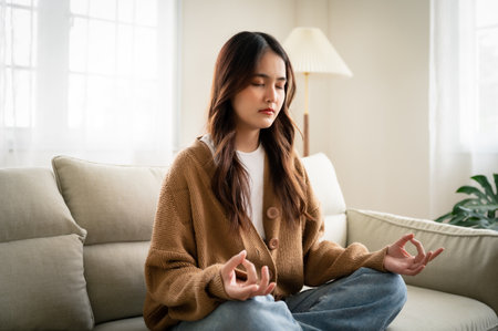 Young asian woman hand meditating and sitting on couch in living room at houseの写真素材