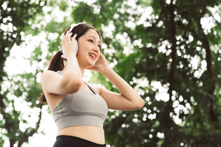 Asian woman trainer is engaged in fitness in public park. Female wearing sport bra and using headphone listening to music. Healthy lifestyle, sports outdoor activities in parkの写真素材