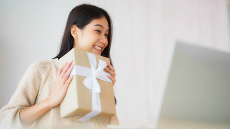 Young asian woman using computer laptop at home. Female showing gift box while on video conversation with friend. Happy birthday, Happy new year, Thanksgivingの写真素材