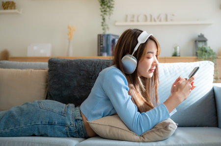 Happy young asian woman relaxing at home. Female smile sitting on sofa and holding mobile smartphone. Girl using video call to friend. Listening to musicの写真素材