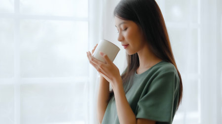 Young asian woman standing beside window and holding mug in bedroom at home, She drinking milk after wake up in the morningの写真素材