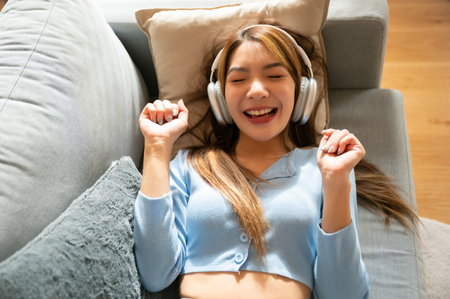 Young asian woman listening to music on couch in living room at home. Happy asia female using mobile smartphone, wearing headset and lying down on sofaの写真素材