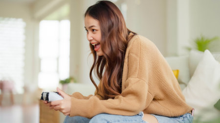 Beautiful young asian woman sitting in living room sofa holding joystick remote control playing...