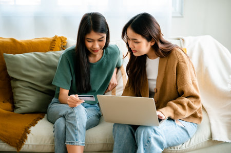 Two asian young woman happy smiling and using computer laptop on couch in living room at homeの写真素材