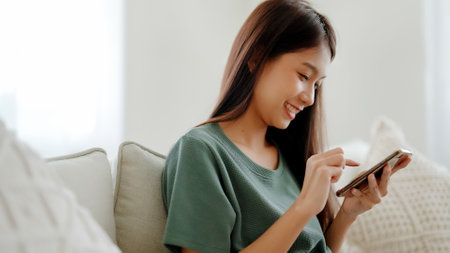 Happy young asian woman relax on comfortable couch at home texting messaging on smartphone, smiling girl use cellphone chatting, browse wireless internet on gadget, shopping online from homeの写真素材