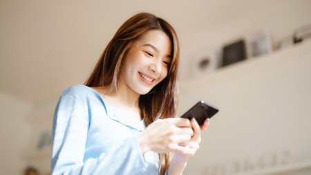Happy young asian woman relax on comfortable couch at home texting messaging on smartphone, smiling girl use cellphone chatting, browse wireless internet on gadget, shopping online from homeの写真素材