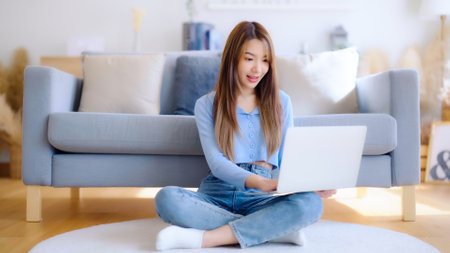 Young asian woman in good spirits working on laptop at home while sitting on the floor close to the couchの写真素材