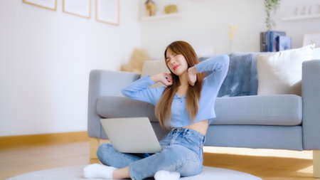 Office syndrome concept. Asian young woman working on laptop while seated on floor next to couch at home, feeling pain in neck and shoulder after working on computer laptop for a long timeの写真素材