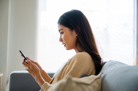 Happy young asian woman relax on comfortable couch at home texting messaging on smartphone, smiling girl use cellphone chatting, browse wireless internet on gadget, shopping online from homeの写真素材
