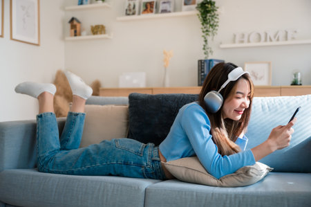 Happy young asian woman relaxing at home. Female smile sitting on sofa and holding mobile smartphone. Girl using video call to friend. Listening to musicの写真素材