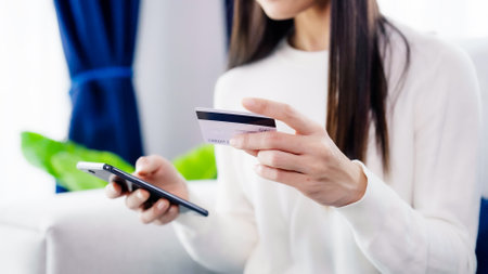Young asian woman sitting on sofa in living room, makes online banking payments through the internet from bank card on smartphone. Shopping online on mobile phone with credit cardの写真素材