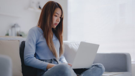 Young asian woman using laptop while seated on couch at homeの写真素材