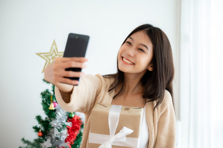 Young asian woman having fun taking selfie and video call talking with her friend. Female showing gift box while on video conversation with friend. Happy new year, Thanksgiving, Merry Christmasの写真素材