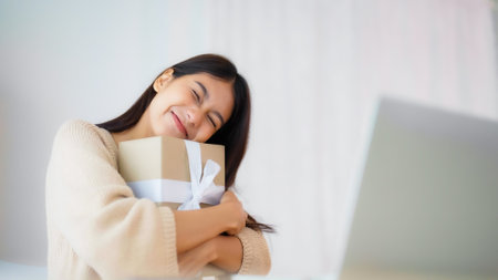 Young asian woman using computer laptop at home. Female showing gift box while on video conversation with friend. Happy birthday, Happy new year, Thanksgivingの写真素材