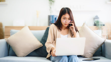 Young asian woman using computer laptop and mobile smartphone while seated on couch at homeの写真素材