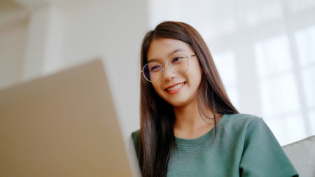 Smiling young asian woman working on laptop at home. Attractive asia female using computer remote studying, watching online education and video callingの写真素材
