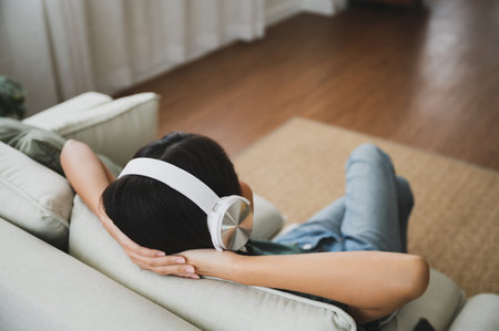 Young asian woman listening to music on couch in living room at home. Happy asia female using mobile smartphone, wearing headset and sitting on sofaの写真素材