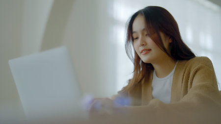 Smiling asian young woman working on laptop at home office. Young asian student using computer remote studying, virtual training, e-learning, watching online education webinar at houseの写真素材