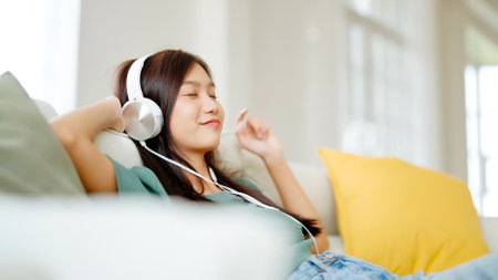 Young asian woman listening to music on couch in living room at home. Happy asia female using mobile smartphone, wearing headset and sitting on sofaの写真素材
