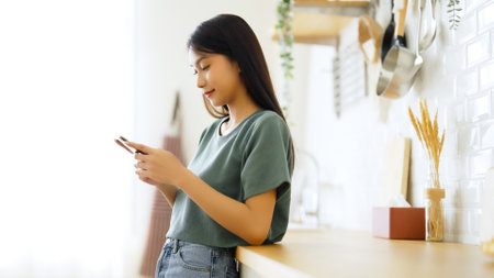 Happy young asian woman relaxing at home. Asia female standing at counter kitchen and using mobile smartphoneの写真素材