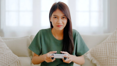 Beautiful young asian woman sitting on sofa and holding joystick playing online video game in living room at houseの写真素材