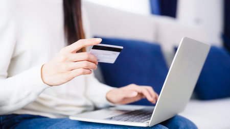 Young asian woman sitting on sofa in living room, makes online banking payments through the internet from bank card on computer laptop. Shopping online on notebook with credit cardの写真素材