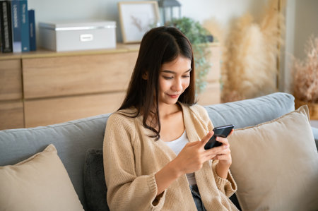Happy young asian woman relax on comfortable couch at home texting messaging on smartphone, smiling girl use cellphone chatting, browse wireless internet on gadget, shopping online from homeの写真素材