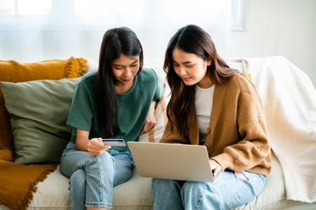 Two asian young woman happy smiling and using computer laptop on couch in living room at homeの写真素材