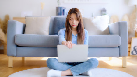 Young asian woman in good spirits working on laptop at home while sitting on the floor close to the couchの写真素材