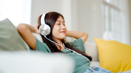 Young asian woman listening to music on couch in living room at home. Happy asia female using mobile smartphone, wearing headset and sitting on sofaの写真素材