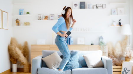 On the sofa in her living room, a young Asian woman dances. Asian woman with a smile relaxing at home, happy and in good mental and physical health, wearing headset and listening to musicの写真素材