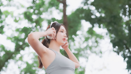 Asian woman trainer is engaged in fitness in public park. Female wearing sport bra and using headphone listening to music. Healthy lifestyle, sports outdoor activities in parkの写真素材