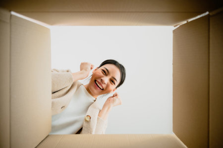 Surprised young asian woman unpacking. Opening carton box and looking inside. Packaging box, delivery service. Human emotions and facial expressionの写真素材