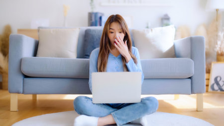Young asian woman in good spirits working on laptop at home while sitting on the floor close to the couch. Excited female winner celebrating success while glancing at notebookの写真素材