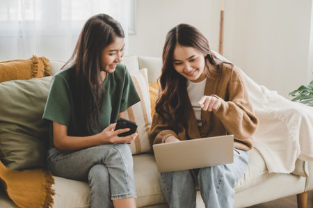 Two asian young woman happy smiling and using computer laptop on couch in living room at homeの写真素材