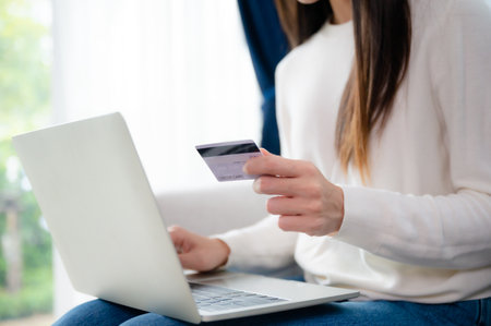 Young asian woman sitting on sofa in living room, makes online banking payments through the internet from bank card on computer laptop. Shopping online on notebook with credit cardの写真素材