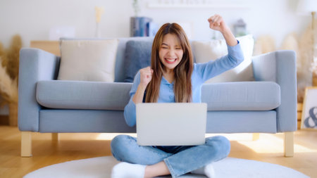 Young asian woman in good spirits working on laptop at home while sitting on the floor close to the couch. Excited female winner celebrating success while glancing at notebookの写真素材