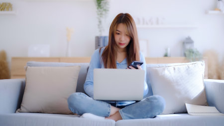 Young asian woman using computer laptop and mobile smartphone while seated on couch at homeの写真素材