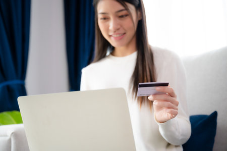 Young asian woman sitting on sofa in living room, makes online banking payments through the internet from bank card on computer laptop. Shopping online on notebook with credit cardの写真素材