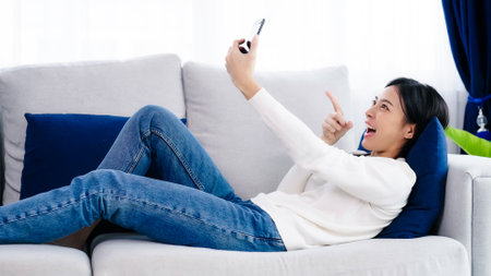 Happy young asian woman relaxing at home. Female smile lying on sofa and holding mobile smartphone. Girl using video call to friendの写真素材