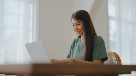 Smiling asian young woman working on laptop at home office. Young asian student using computer remote studying, virtual training, e-learning, watching online education webinar at houseの写真素材