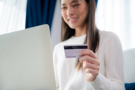 Young asian woman sitting on sofa in living room, makes online banking payments through the internet from bank card on computer laptop. Shopping online on notebook with credit cardの写真素材