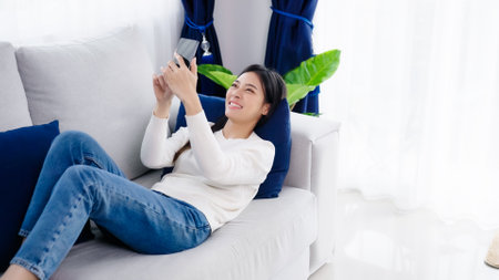 Happy young asian woman relaxing at home. Female smile lying on sofa and holding mobile smartphone. Girl using video call to friendの写真素材