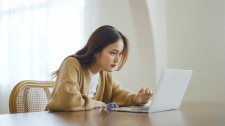 Smiling asian young woman working on laptop at home office. Young asian student using computer remote studying, virtual training, e-learning, watching online education webinar at houseの写真素材